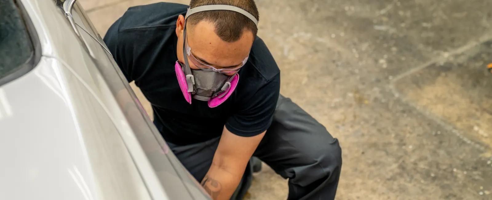 Mechanic wearing protective mask inspecting or repairing a white car in a garage with concrete floor