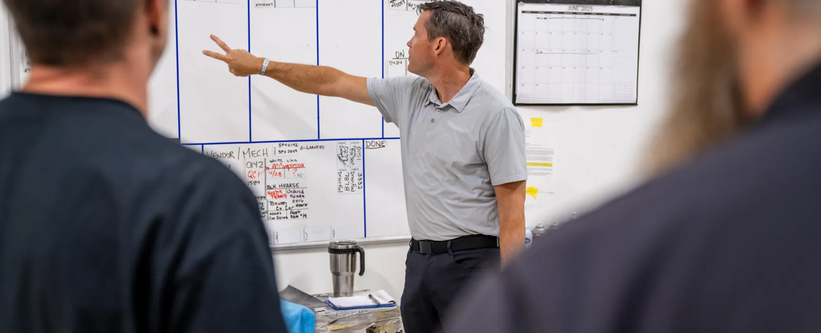 Man pointing to a whiteboard with work stages while two colleagues listen in a workshop or office setting