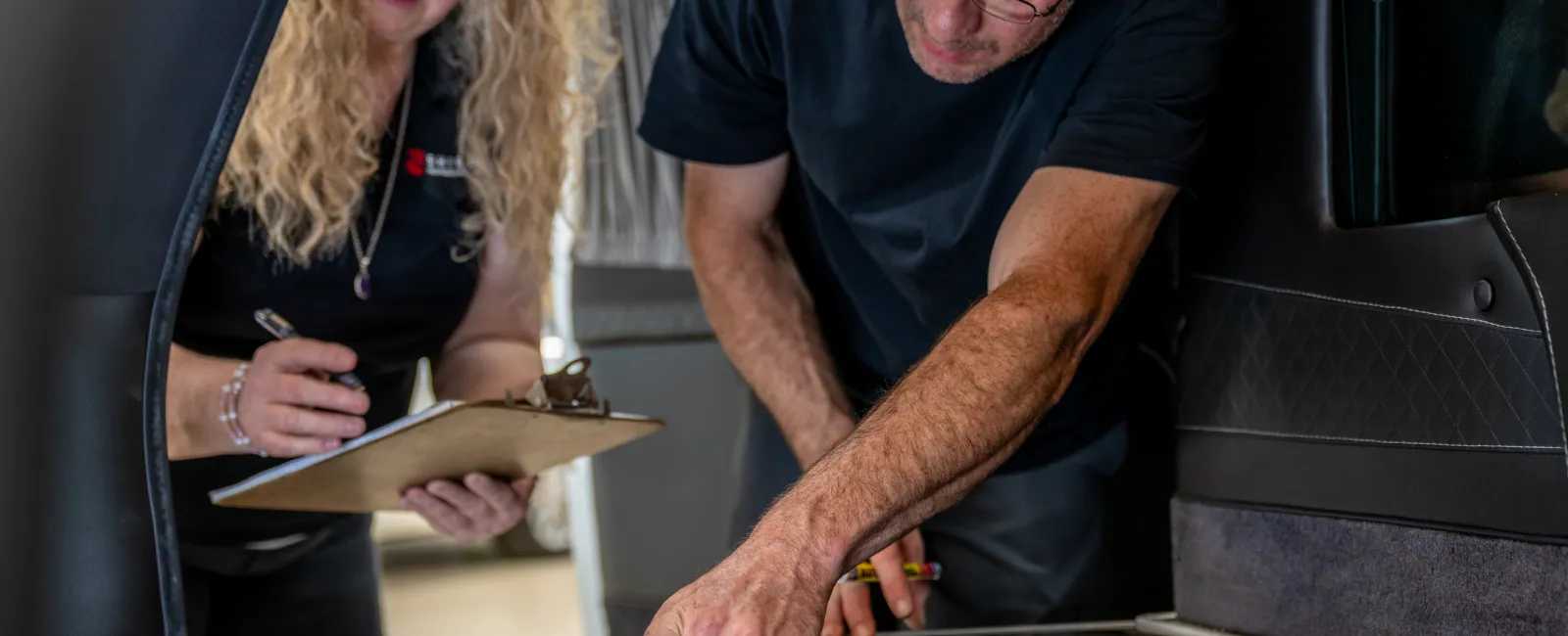 Two workers inspecting and measuring a vehicle floor with tools while one takes notes on a clipboard in a workshop.