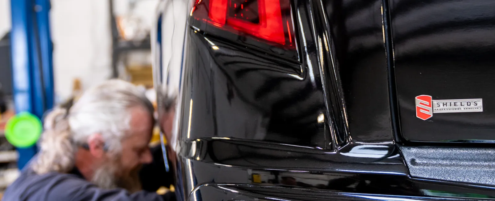 Mechanic working on the rear left side of a black luxury car in a workshop garage setting.