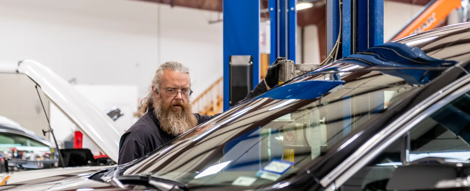 Mechanic with beard inspecting a black car inside an automotive repair shop with lifted vehicles.