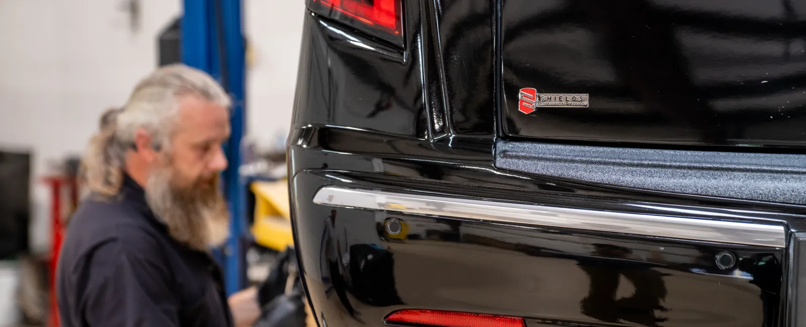 Close-up of a black luxury car's rear bumper with a mechanic working in the blurred background in a garage.
