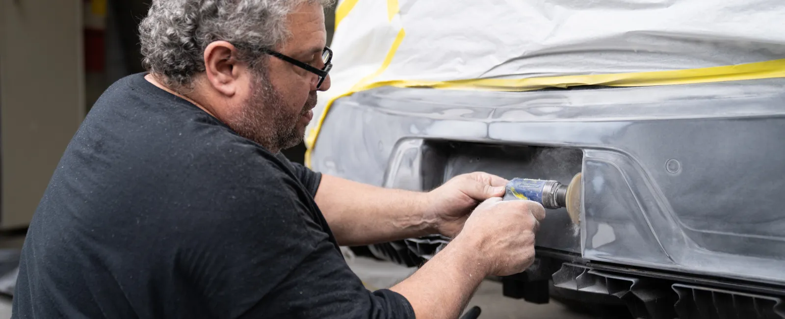 Man using a power tool to sand the rear bumper of a car prepped with masking tape for painting