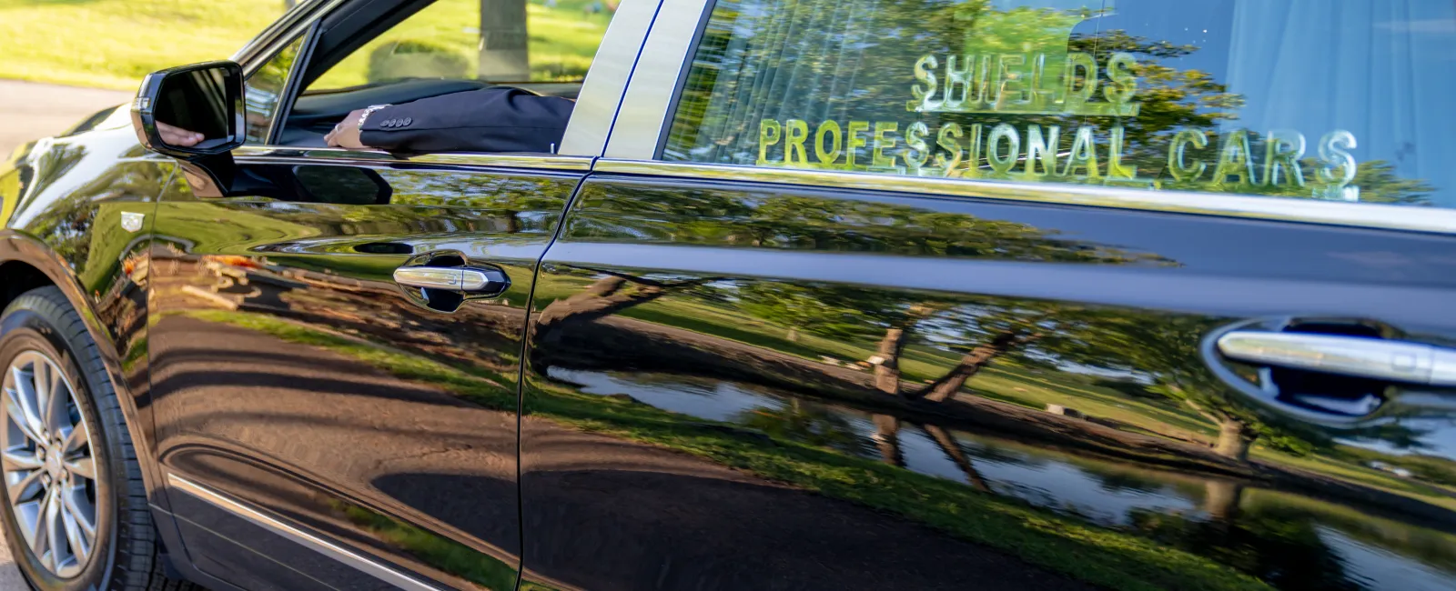 Black luxury sedan with professional chauffeur driving past green park landscape on a sunny day