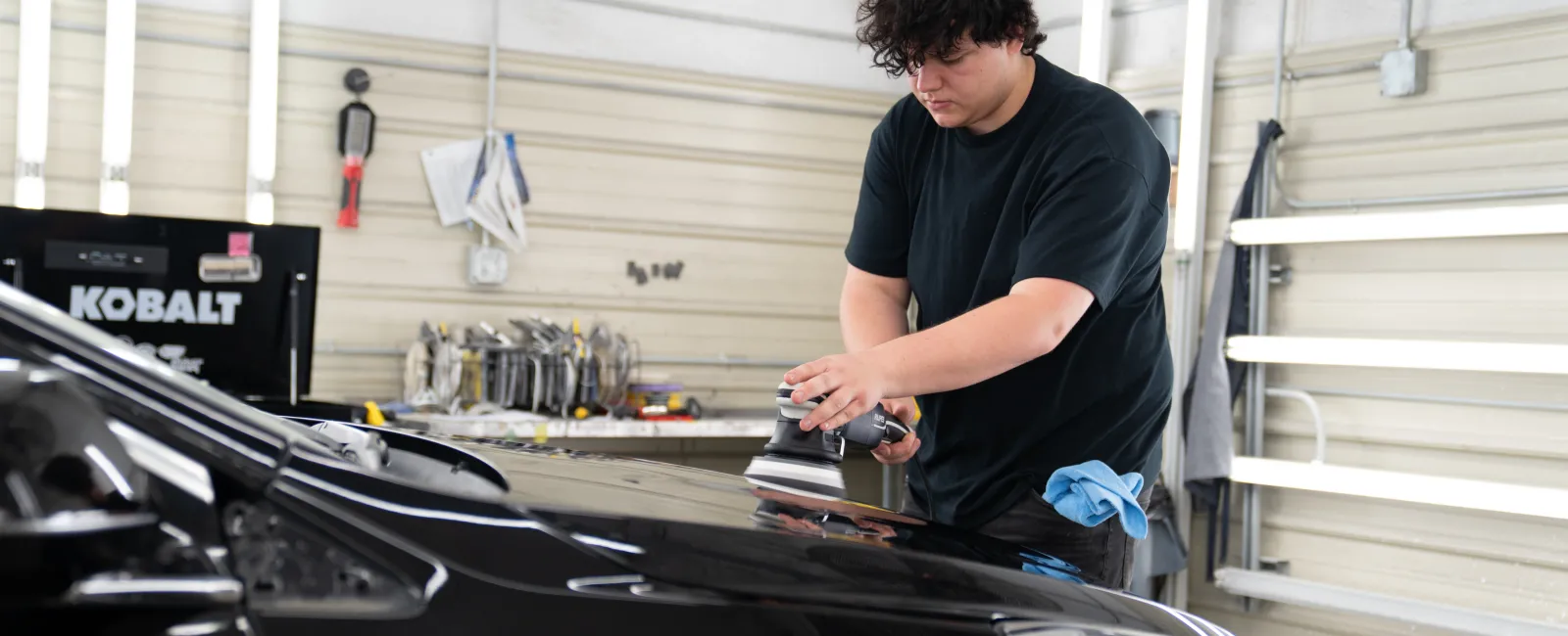 Person polishing the black hood of a car inside a well-lit workshop with tools and equipment in the background