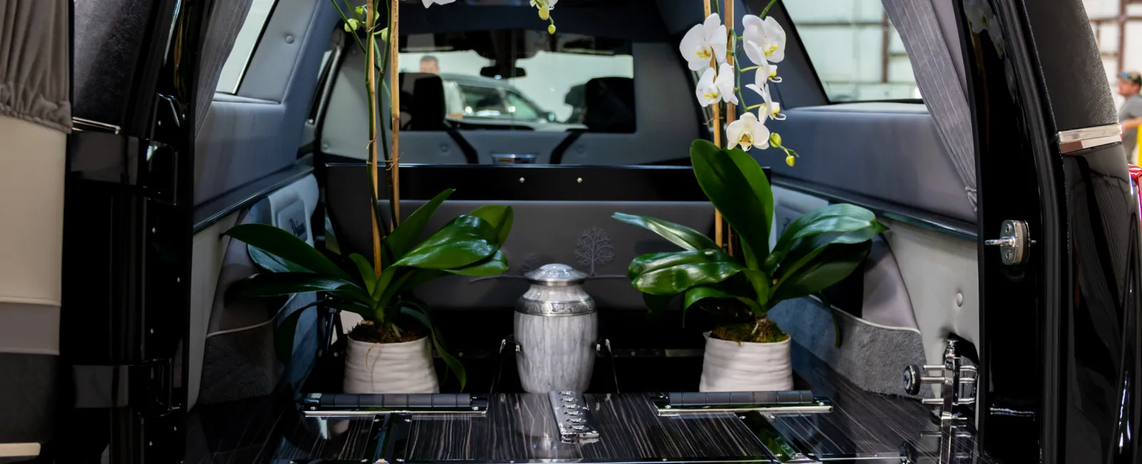 Rear view of a black hearse with open back door showing an urn and white orchids inside in a warehouse