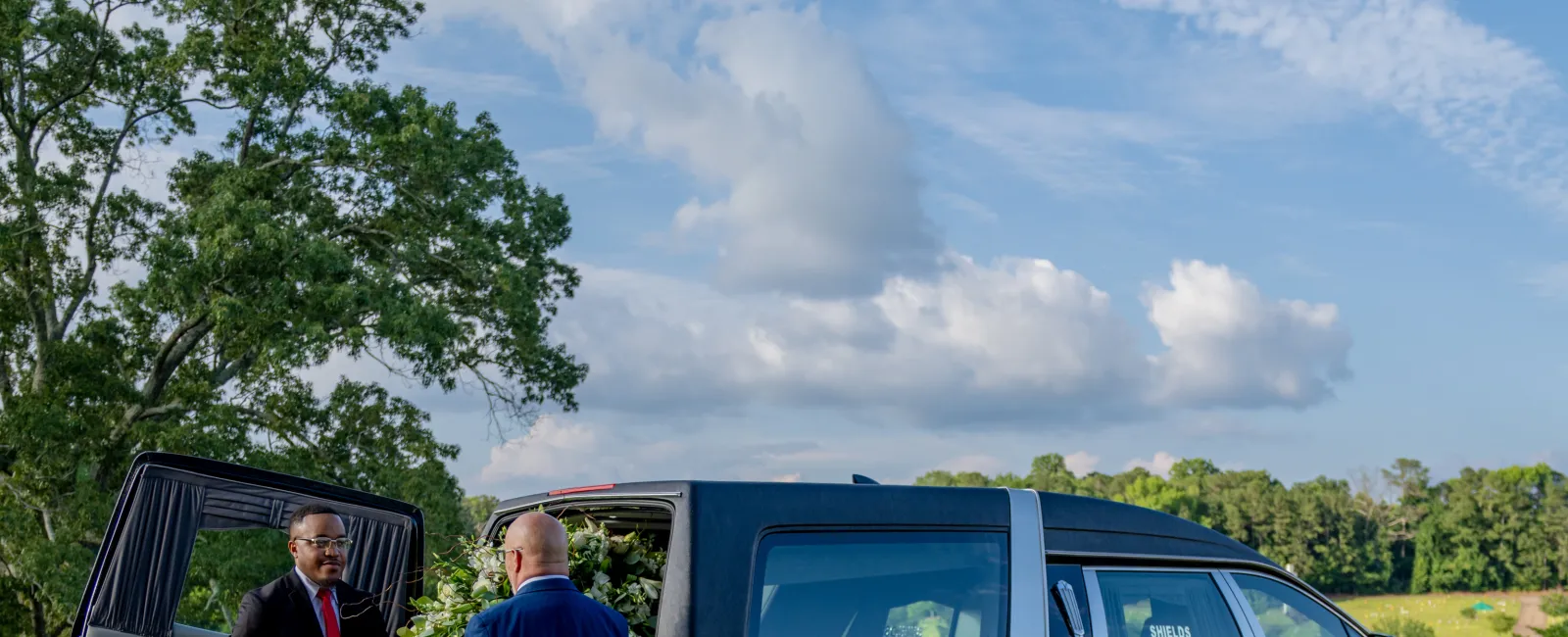 Two men carrying a wooden coffin from a black hearse on a sunny day with trees and blue sky background