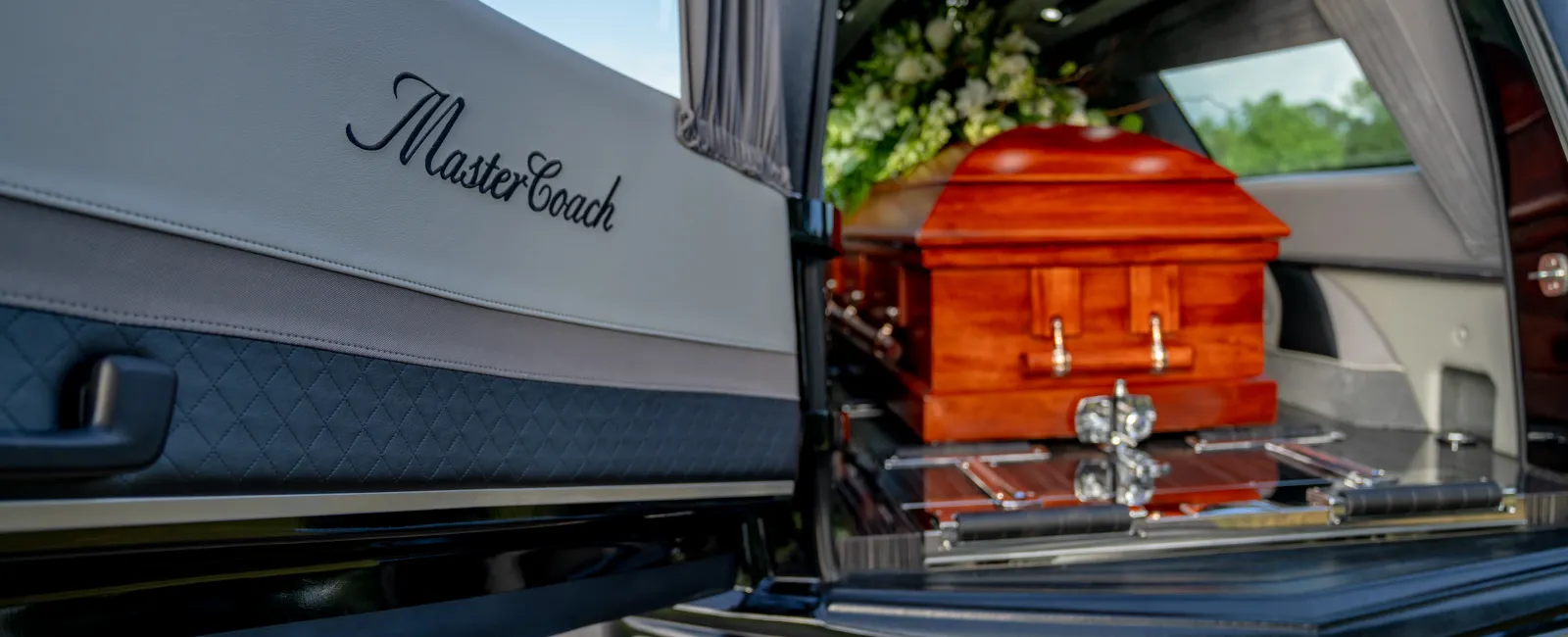 Open hearse with MasterCoach logo on door and wooden casket inside surrounded by floral arrangement