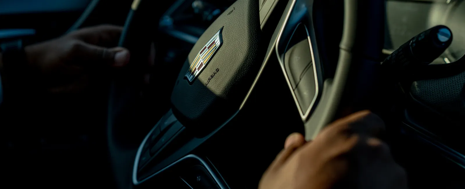 Close-up of hands gripping a Cadillac steering wheel inside a dark car interior with natural light highlighting the logo