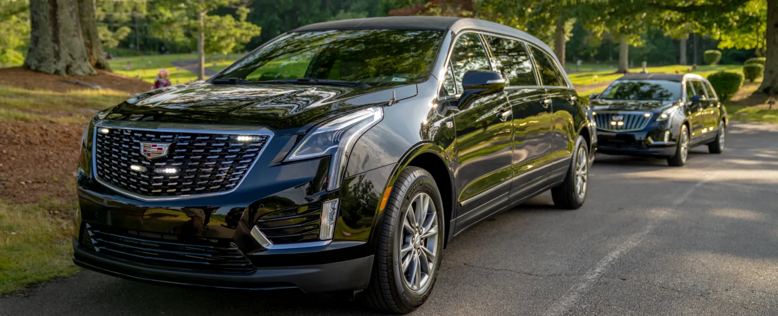 Two black Cadillac SUVs parked on a tree-lined road with sunlight filtering through the leaves
