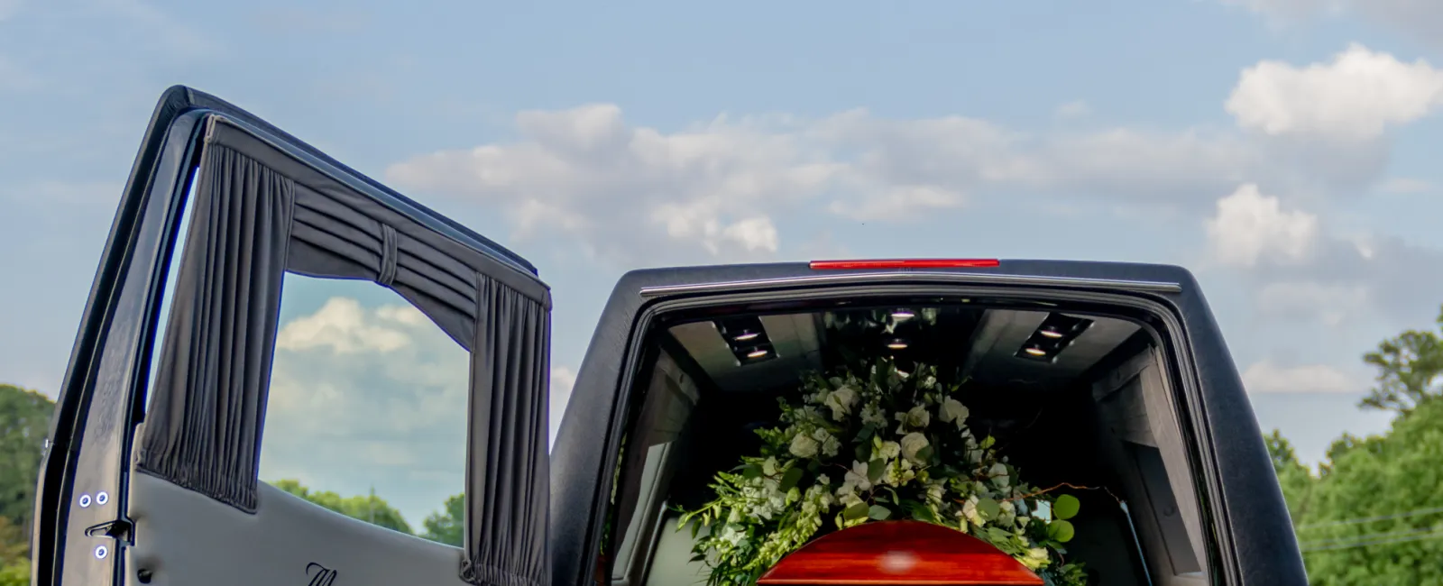 Black hearse with open rear door carrying a wooden coffin decorated with white flowers on a road under blue sky