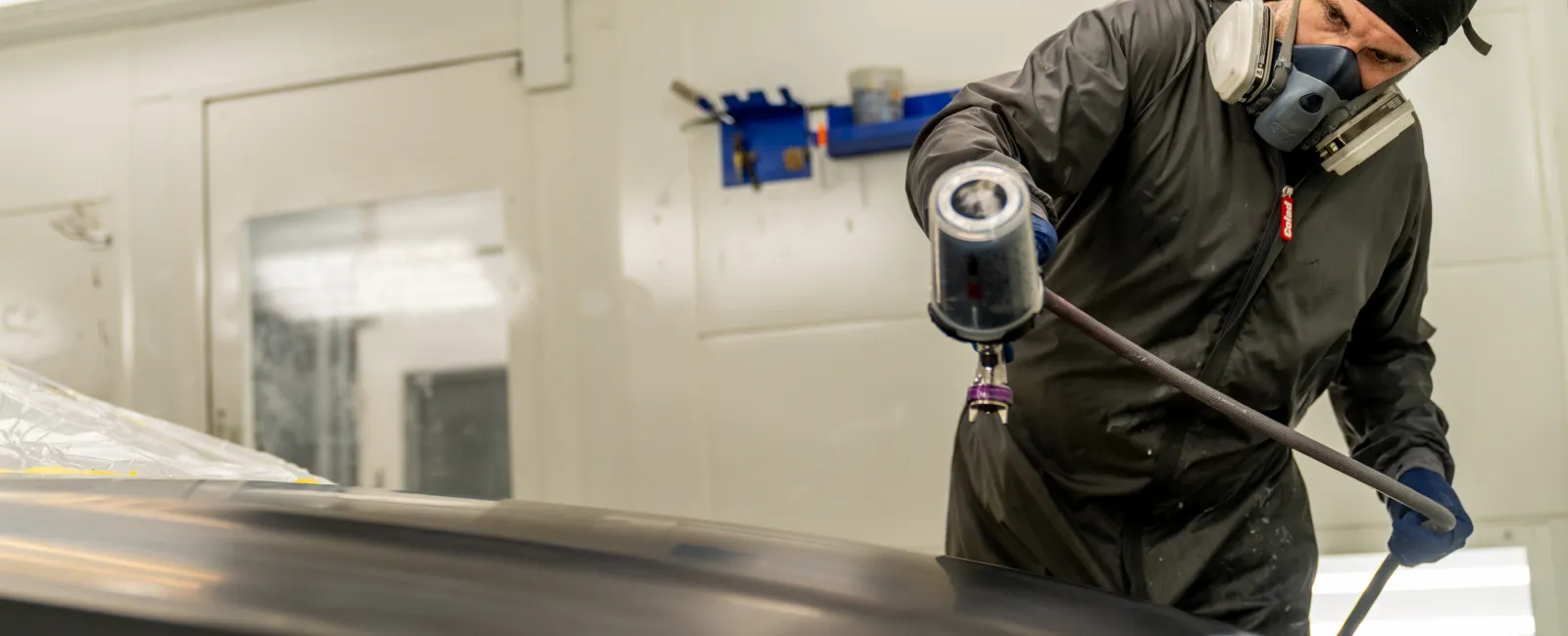Worker in protective gear spraying paint on a car hood in a well-lit automotive paint booth