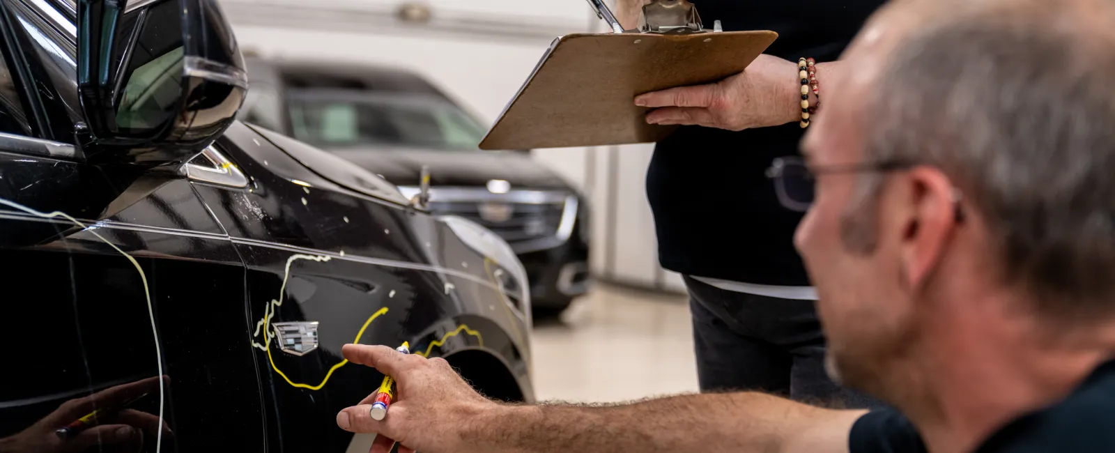 Mechanics inspecting and marking scratches on a black car in an auto repair shop with clipboard notes.
