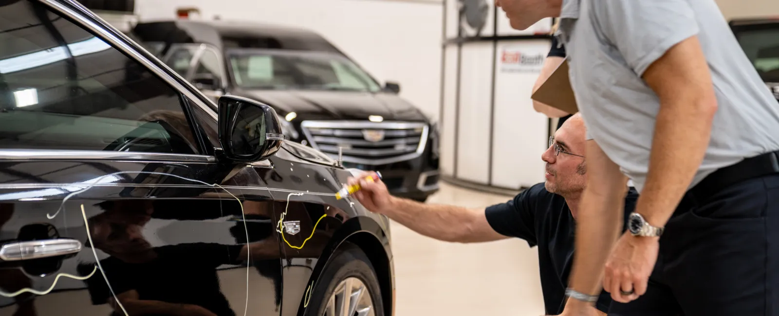 Two men inspecting and marking dents on a black Cadillac car inside a repair workshop with other vehicles in the background