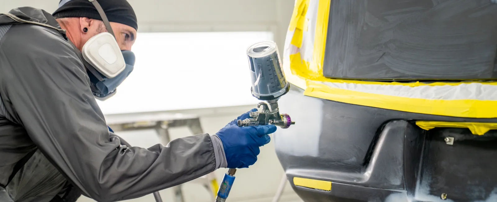 Man wearing protective gear spray painting a car bumper in a professional auto body repair shop