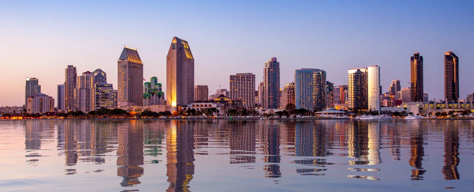 San Diego city skyline at sunset with buildings reflecting on calm harbor water and clear blue sky.