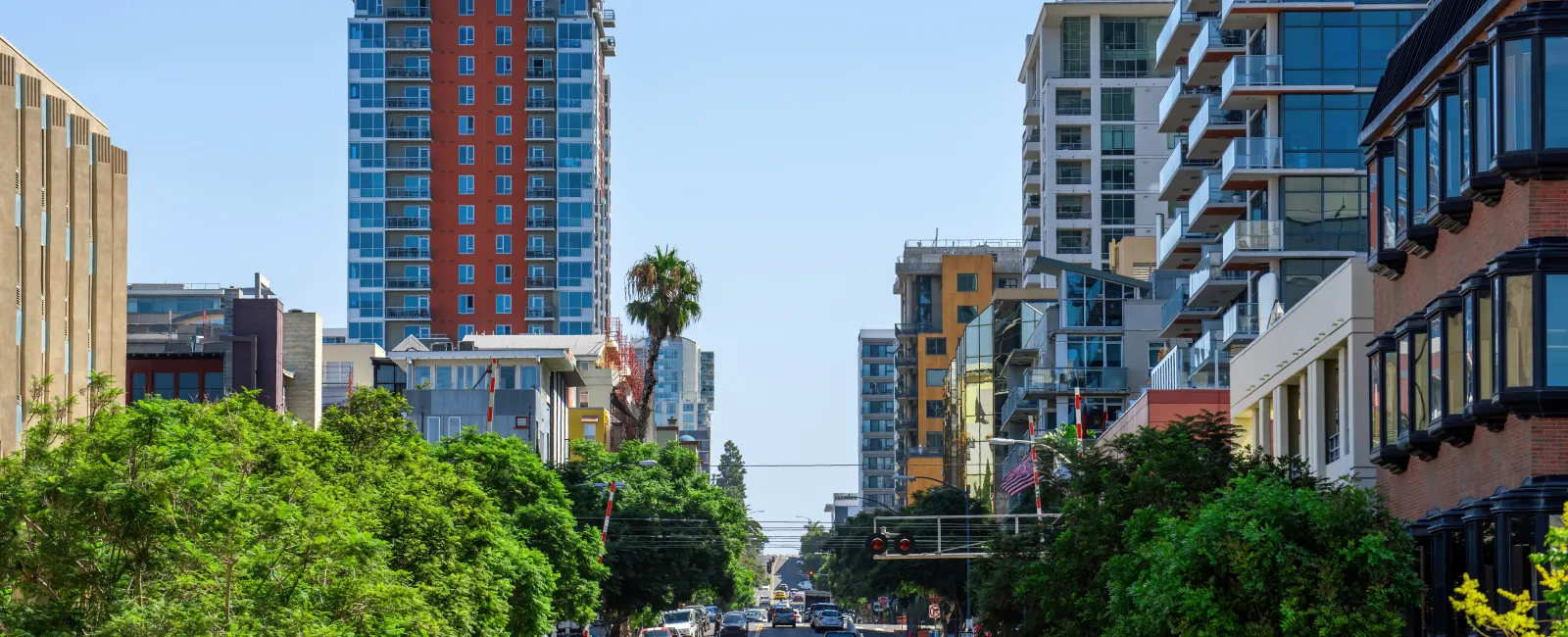 Urban street view with tall buildings, green trees lining the road, and cars parked on both sides under clear blue sky