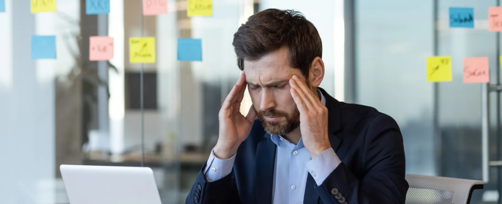 Stressed businessman in suit massaging temples working on laptop in modern office with sticky notes on glass wall.