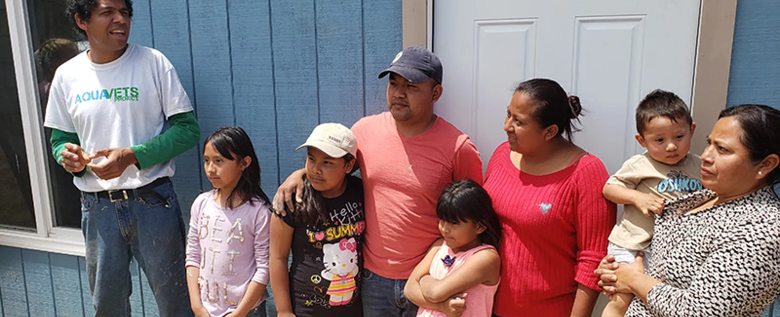 Family of seven standing outside a blue house with a white door on a sunny day.