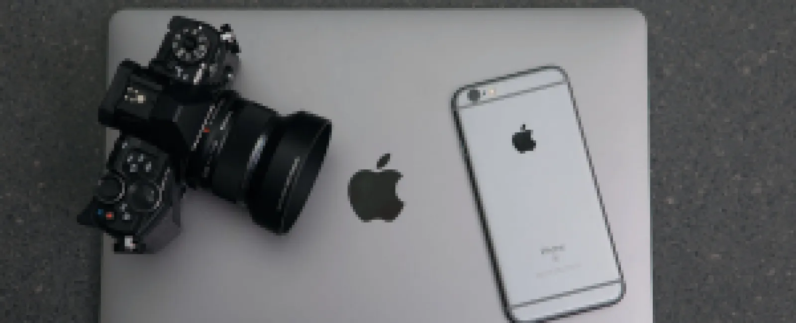 Professional camera and iPhone resting on a closed Apple MacBook on a dark textured surface