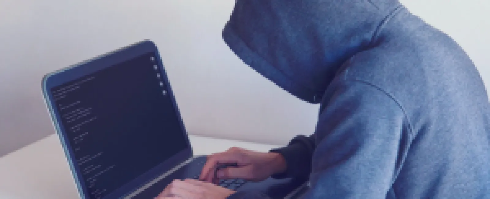 Person in gray hoodie typing on laptop with code visible on screen in a dimly lit room.