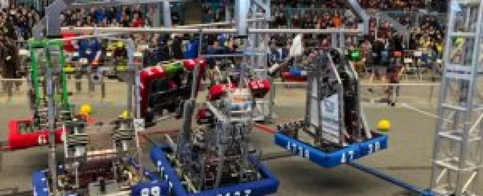 Three numbered competition robots suspended mid-air during a robotics event with spectators in the background.