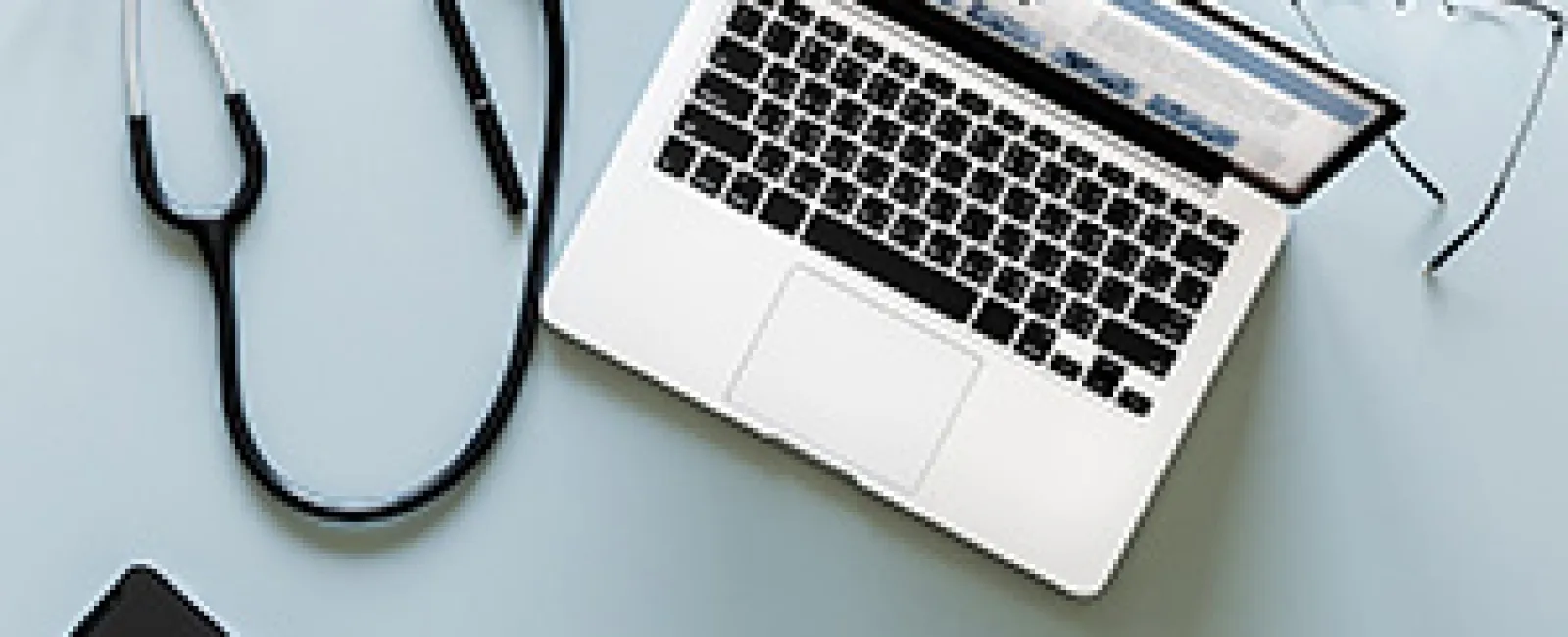 Laptop, stethoscope, eyeglasses, and smartphone on a light surface representing telemedicine setup