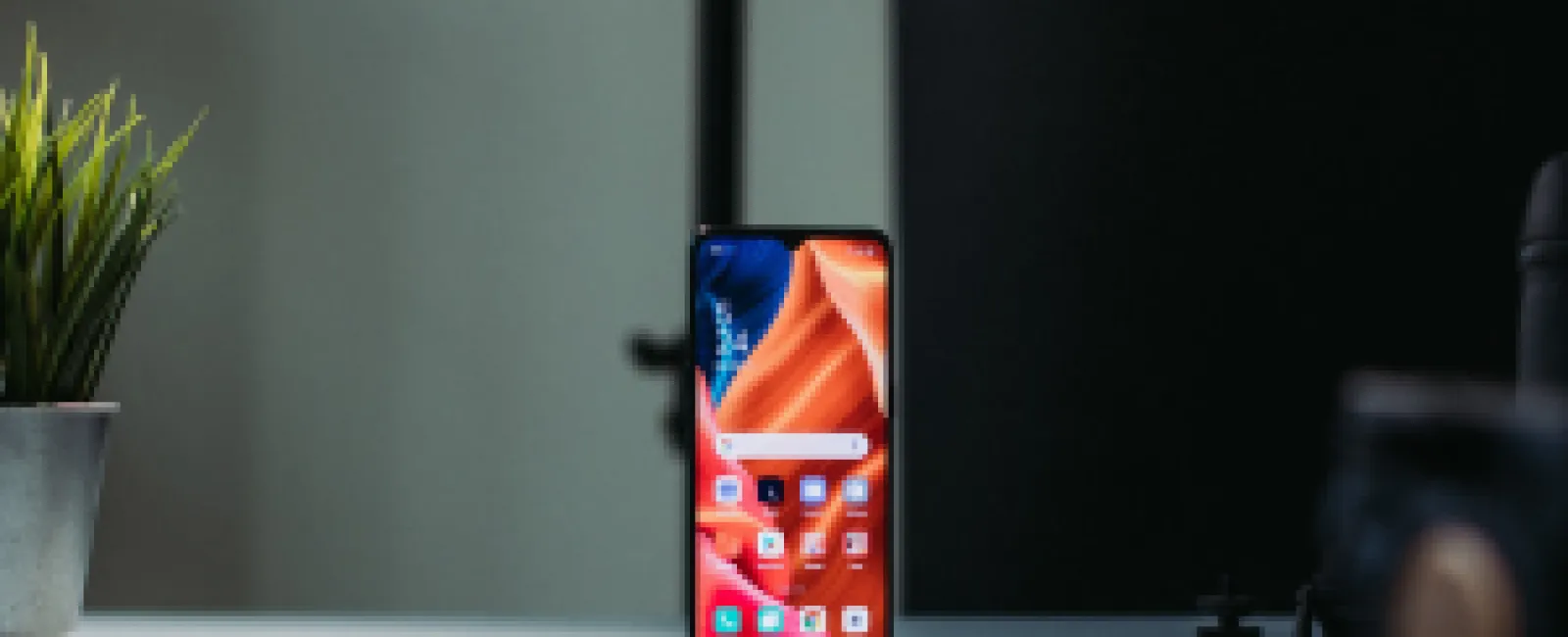 Smartphone with colorful display standing on a white desk next to a potted plant and black background.