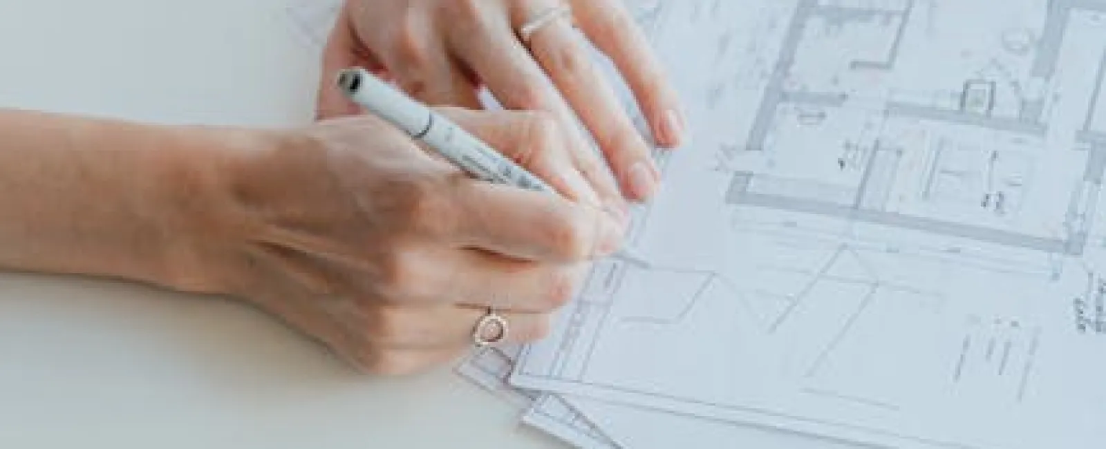 Person drawing architectural floor plans with pen on paper at a desk with sketches and phone nearby