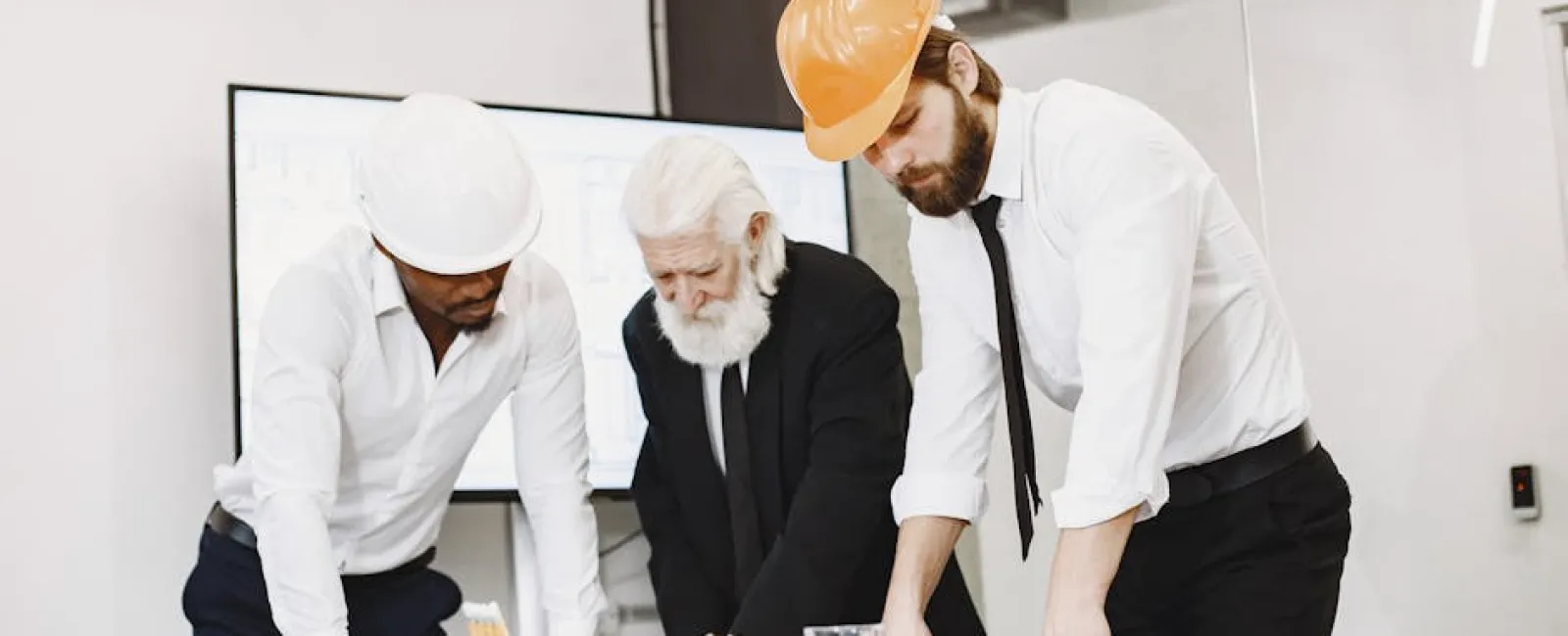 Three engineers in helmets reviewing blueprints and project plans together in a modern office setting.