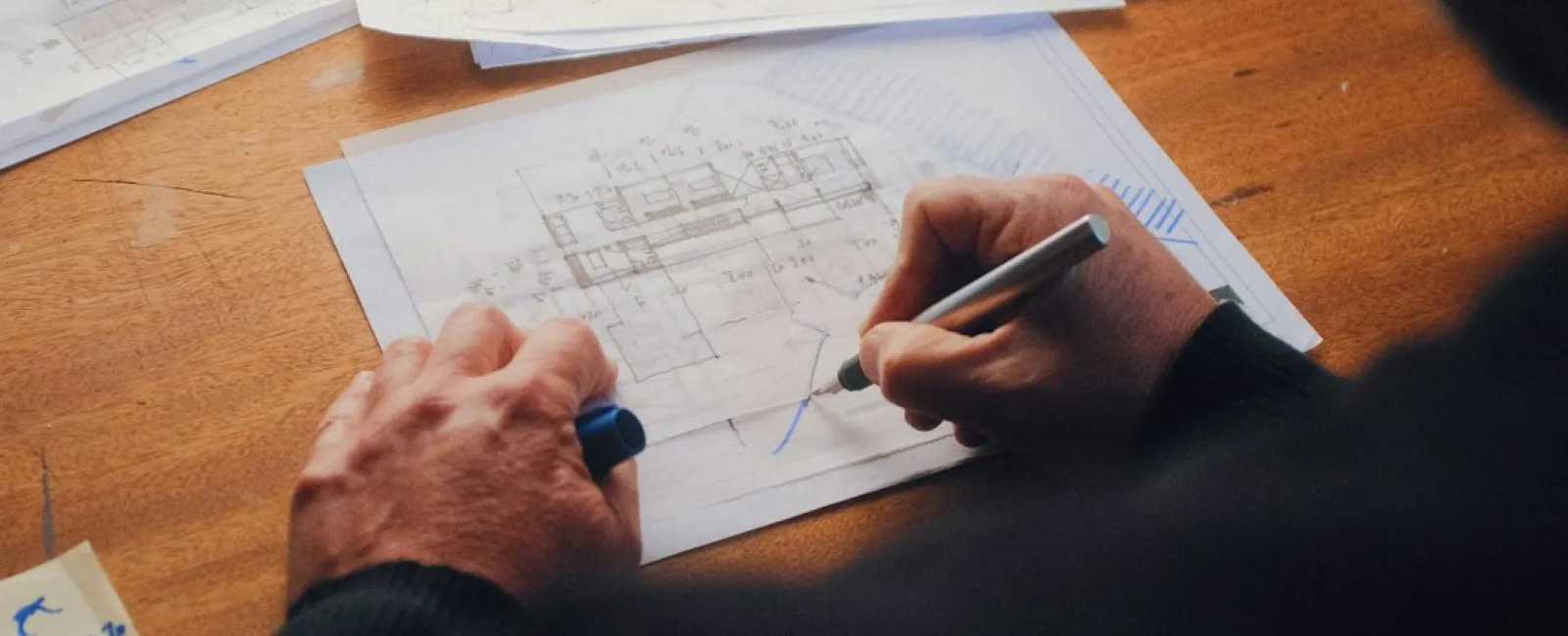 Person sketching architectural plans on paper at a wooden desk with colored pens in the background