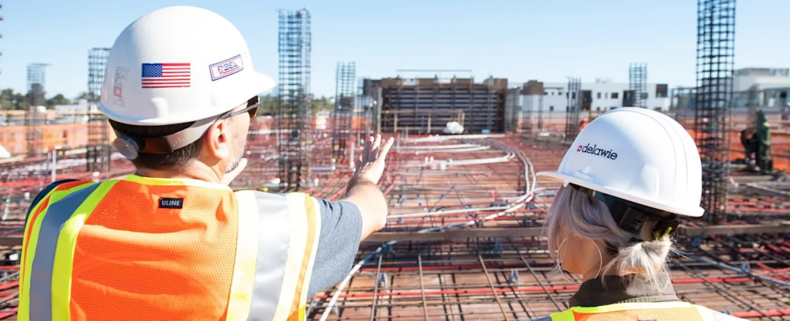 Two construction workers in hard hats and reflective vests inspecting a building site with steel rebar framework.