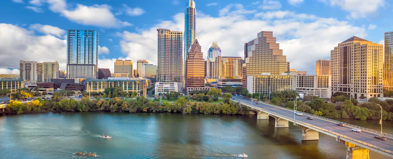 Austin city skyline with tall buildings, a bridge over the river, and boats rowing on the water under a blue sky.