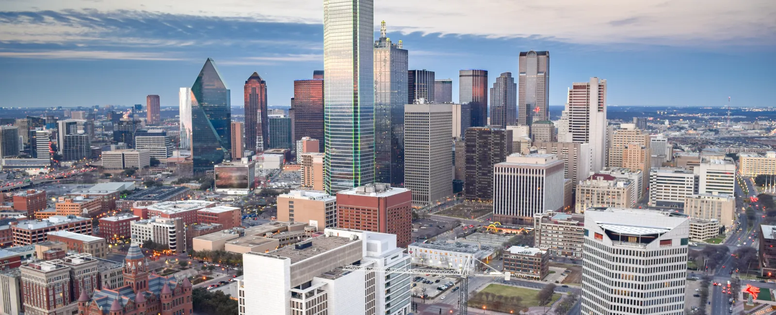 Aerial view of downtown Dallas with tall skyscrapers and city streets at sunset under a blue sky.