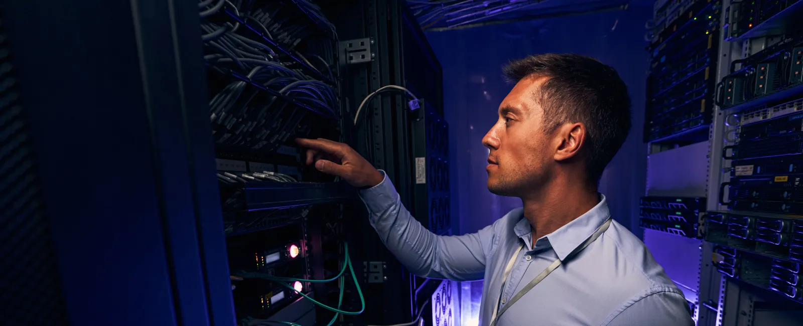 IT technician working on server racks with laptop in dimly lit data center room with blue lighting.