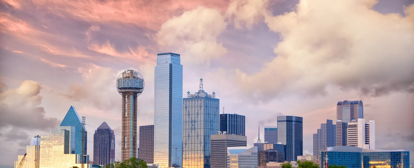 Dallas city skyline at sunset with iconic Reunion Tower and reflective skyscrapers under colorful clouds.