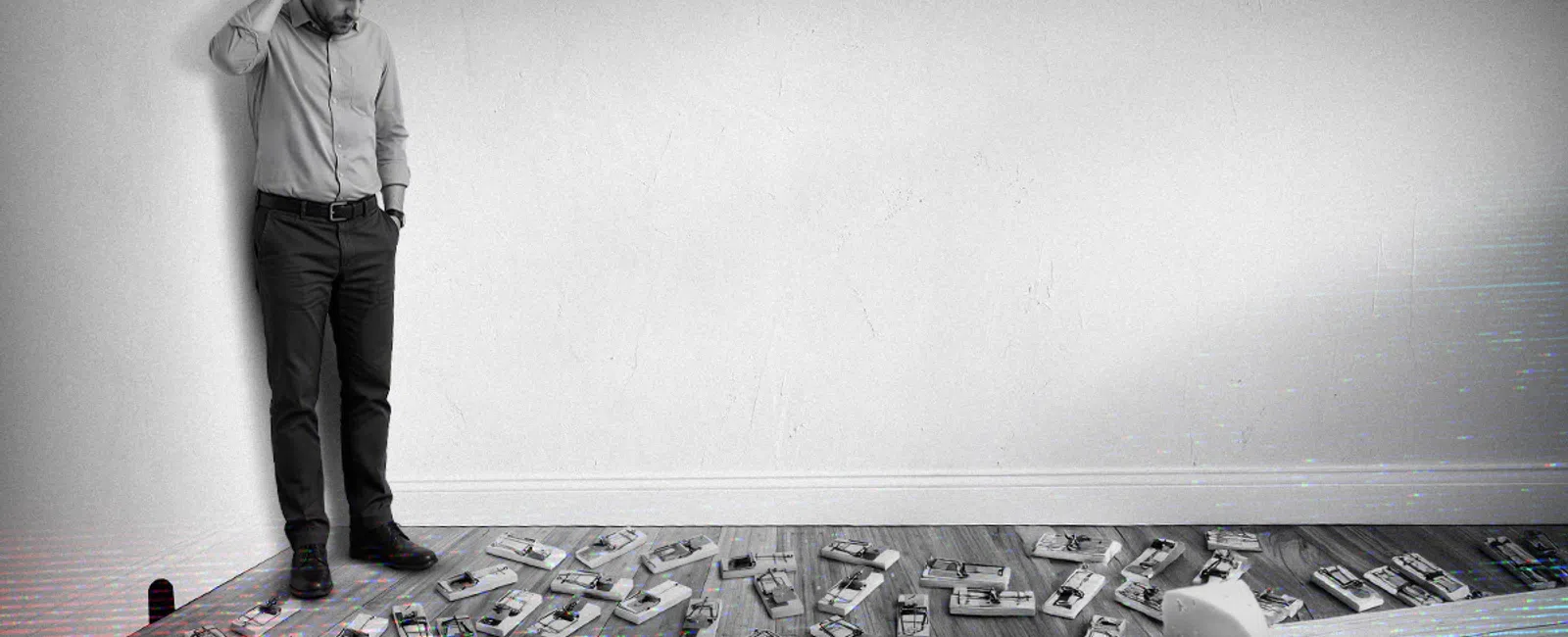 Man in business attire standing confused by a floor filled with numerous mousetraps in a minimalist room.