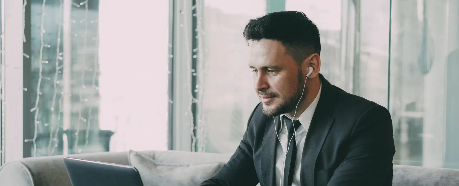 Man in business suit working on laptop with earphones at cafe table with coffee cup and notebook.