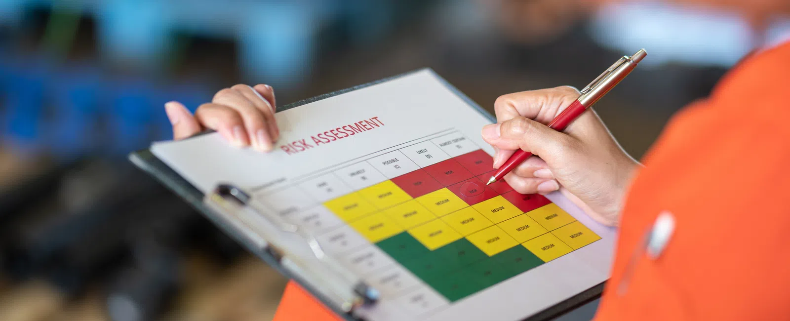 Person in orange holding clipboard with color-coded risk assessment chart and marking with red pen.