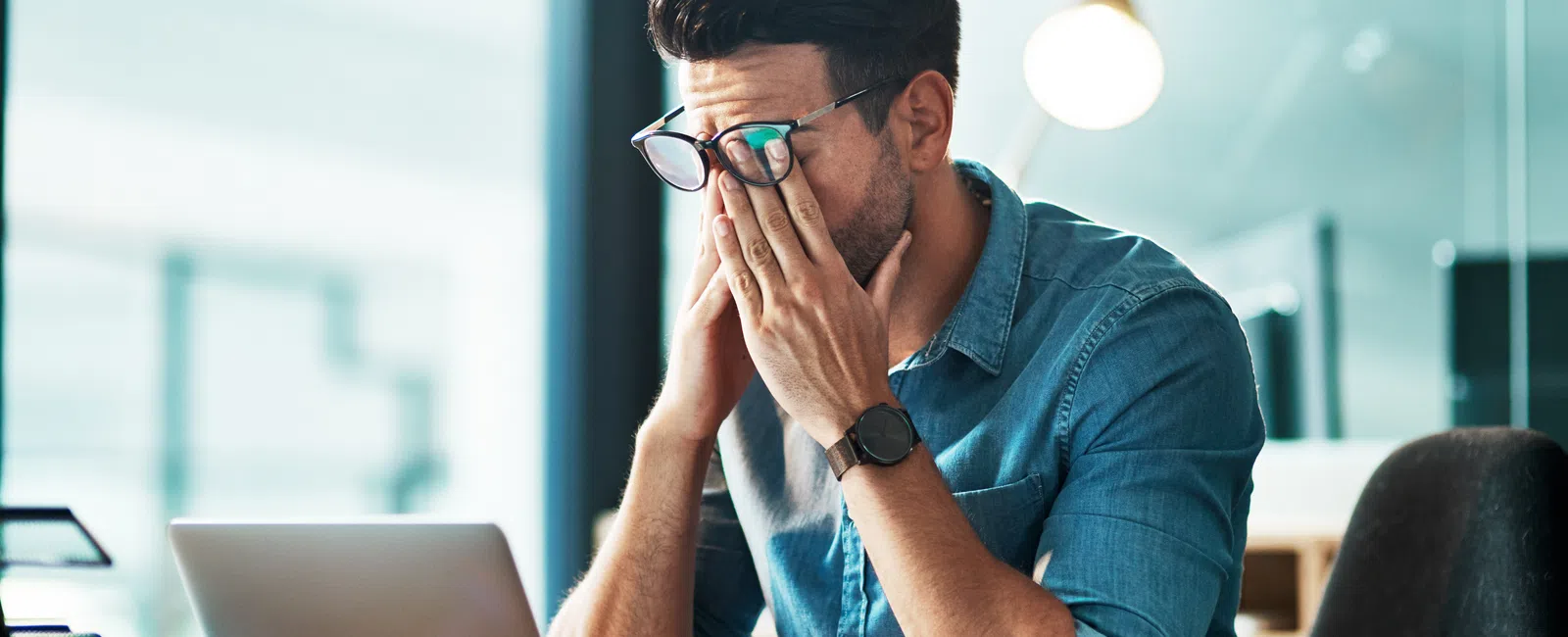 Man in blue shirt rubbing tired eyes sitting at desk with laptop in modern office setting