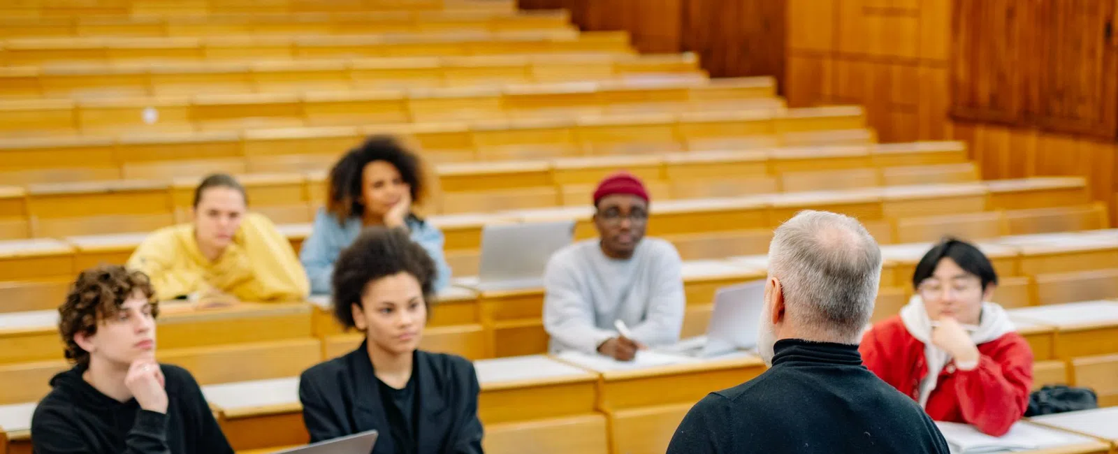 Professor lecturing to attentive diverse students in a wooden-tiered university classroom with laptops open.