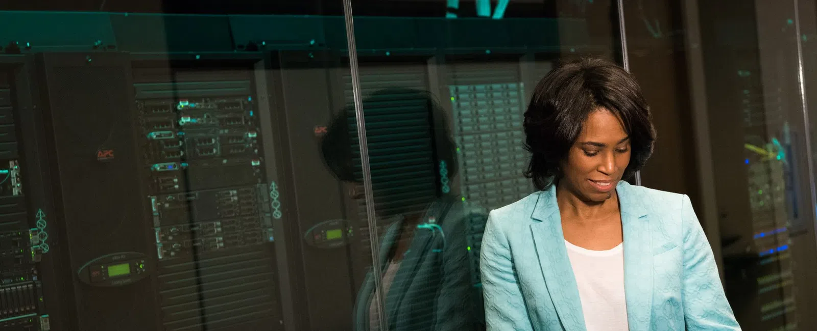 Professional woman in teal blazer using tablet in data center with server racks and glass wall reflections