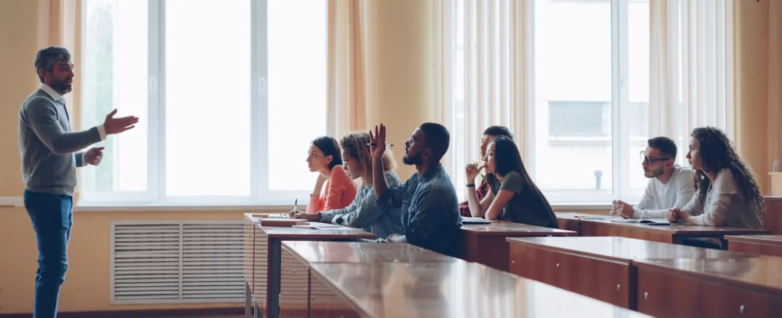 University professor teaching diverse students in a classroom with large windows and wooden desks