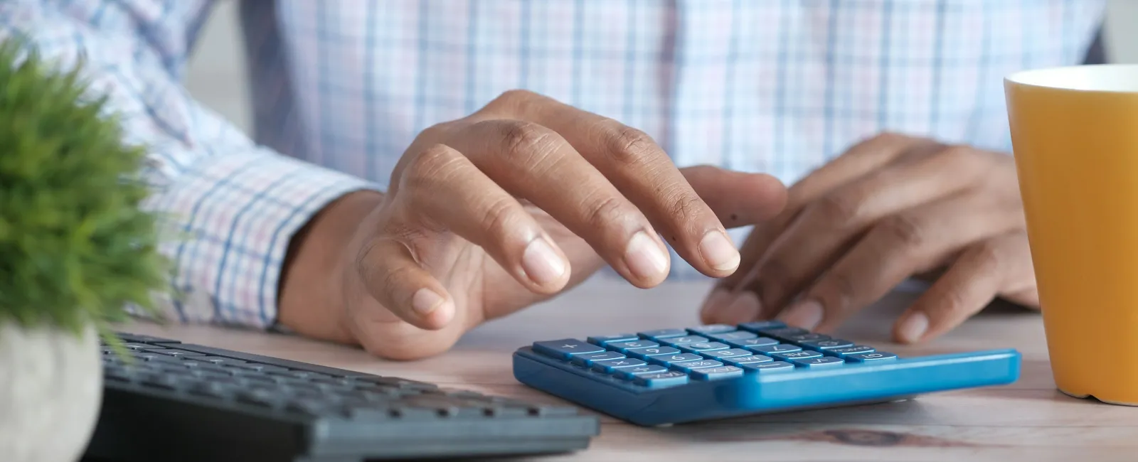 Person using a blue calculator on a wooden desk with a keyboard, plant, and yellow cup nearby