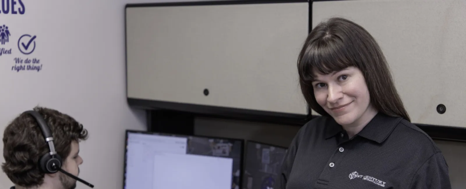 Woman holding a notepad smiling. Man behind her sitting at a desk with two monitors and a headset. Both are employees of Next Century Tech
