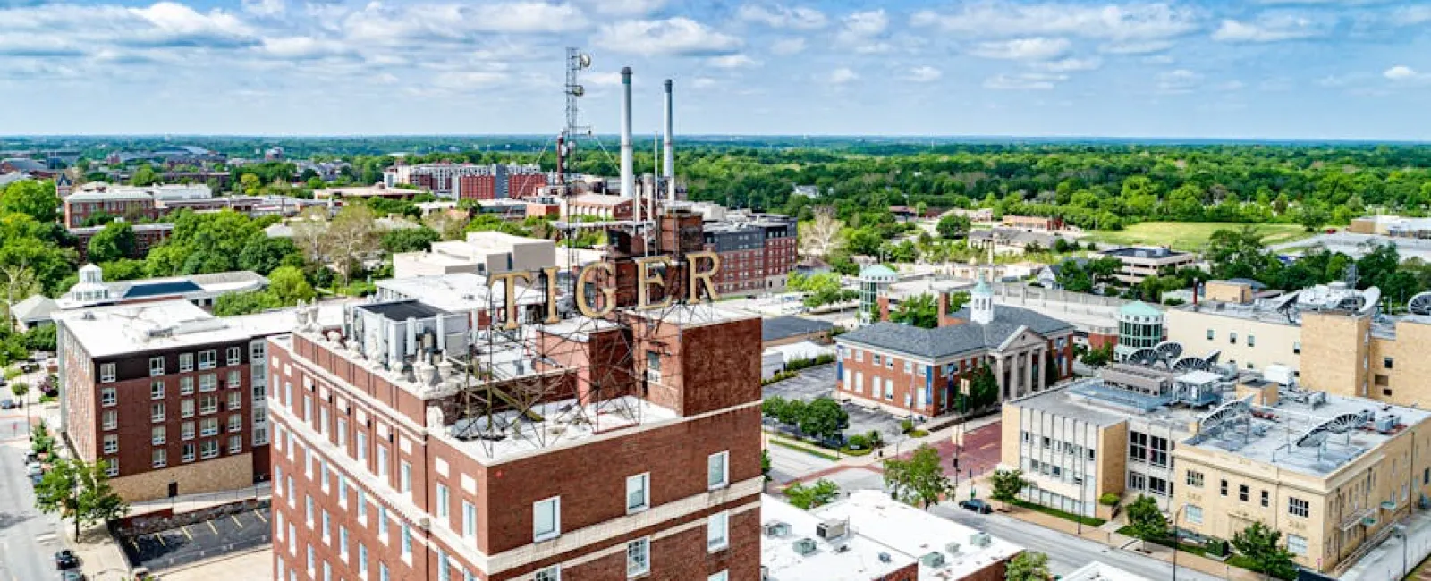 Aerial view of downtown with Tiger sign atop a brick building under a blue sky with clouds in a green city landscape.