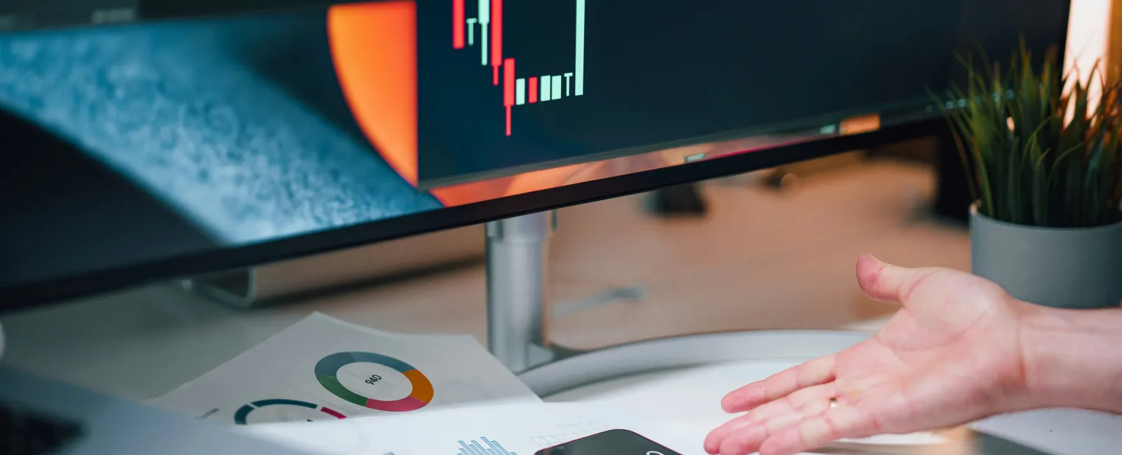 Desk setup with financial charts, calculator, keyboard, and hand gesturing towards stock market data on screen.