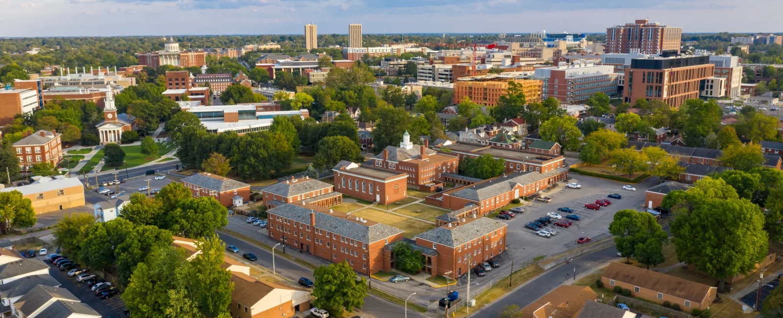 Aerial view of a cityscape showing a mix of residential and commercial buildings with green trees and cloudy sky.