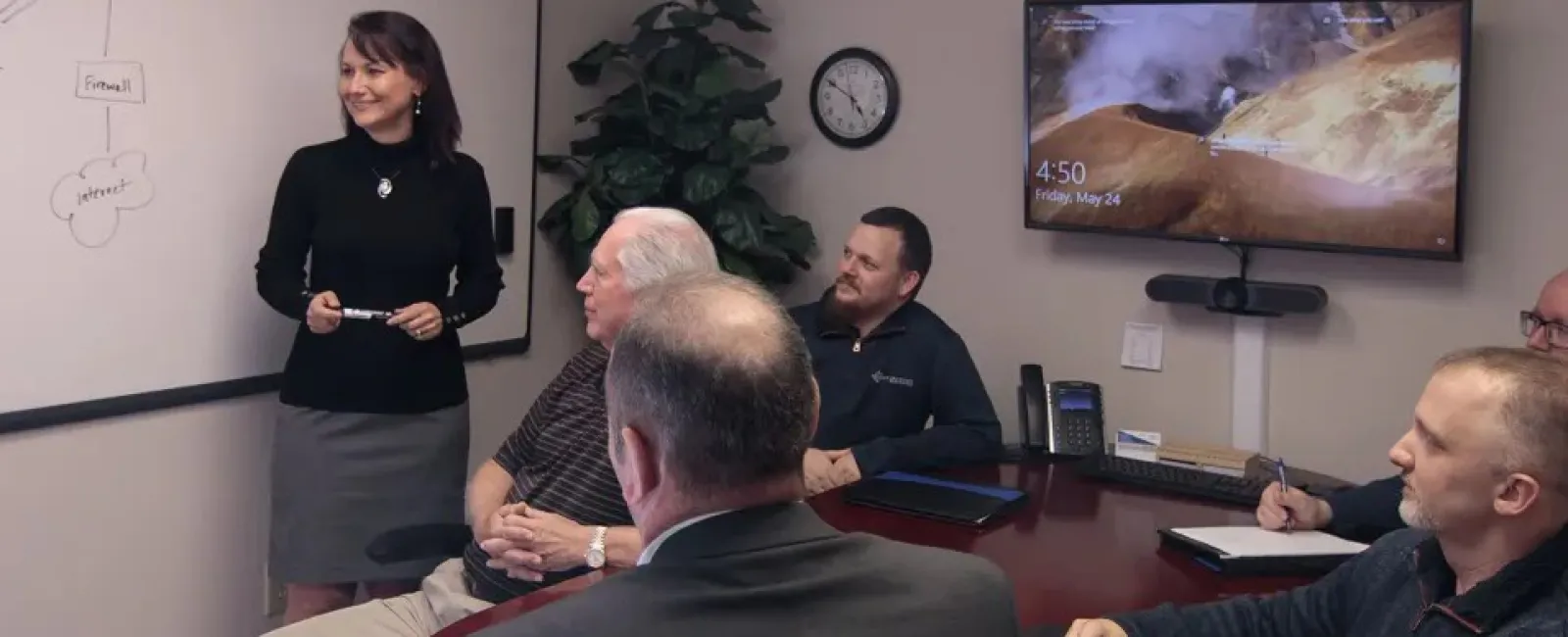 A woman standing at a whiteboard in a conference room. Five men are sitting around the table.