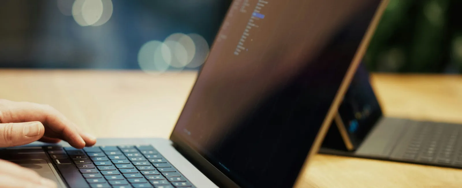 Hands typing on a laptop keyboard displaying code in a dimly lit workspace with blurred background bokeh.